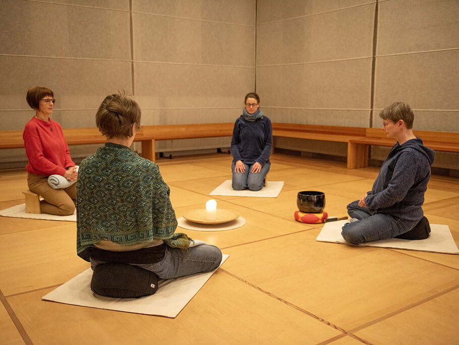 Meditationsgruppe-aspect-ratio-4-3 Vier Frauen knien im Kreis auf dem Boden, haben die Augen geschlossen und meditieren. In der Mitte ihres Sitzkreises steht eine brennende Kerze. Vor einer Fra steht eine Klangschale.